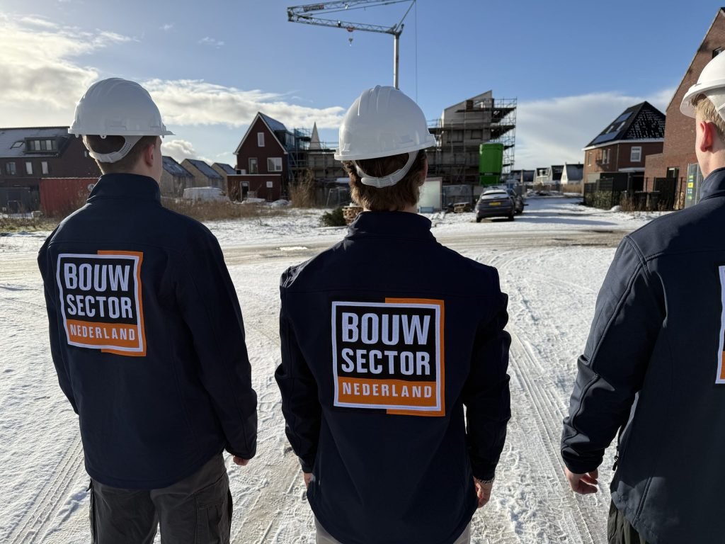Drie vakmensen van Bouwsector Nederland met witte helm, op de rug gezien, kijken naar huizen in aanbouw met een bouwkraan erboven